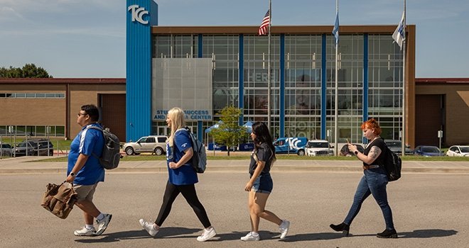 Four 91Ӱ True Blue Lead Students walk across the parking lot in front of southeast campus building.