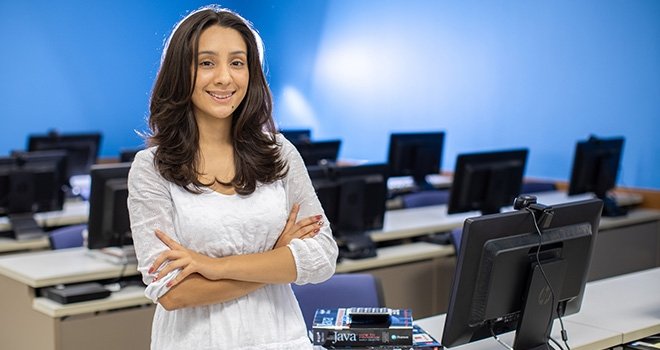 Young woman smiles with arms crossed, standing in a computer lab at 91Ӱ.