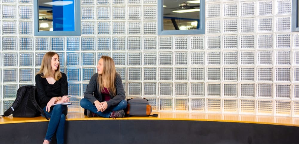 Two 91Ӱ students chatting on a bench in front of a wall of glass bricks